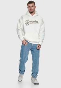 Man wearing white hoodie with "Dropsize" text, blue jeans, and white sneakers, standing against a plain light gray background.