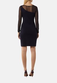 Dopasowana granatowa sukienka z długimi przezroczystymi paskowanymi rękawami, wygiętym dekoltem i gładkim, jednolitym materiałem. Noszona z wysokimi obcasami.