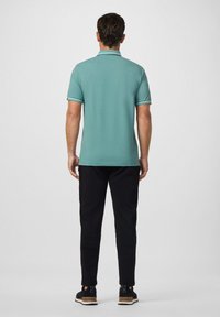 Muž stojící zády k fotoaparátu, oblečený v tyrkysovém polo tričku, černých kalhotách a černých teniskách s hnědými podrážkami na jednobarevném pozadí.