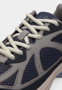 Κοντινή λήψη αθλητικού παπουτσιού σε μπλε ναυτικό και γκρι χρώμα με εκρού διασταυρούμενα κορδόνια και πάνελ από πλεκτό ύφασμα.