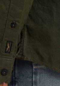 Camisa verde oliva con una tela texturizada, botones negros y una pequeña etiqueta negra con detalles en dorado en el dobladillo.