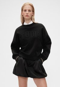 Sudadera negra con texto en relieve "STUDIO", corte relajado, escote redondo, puños acanalados, combinada con pantalones cortos de satén negro.