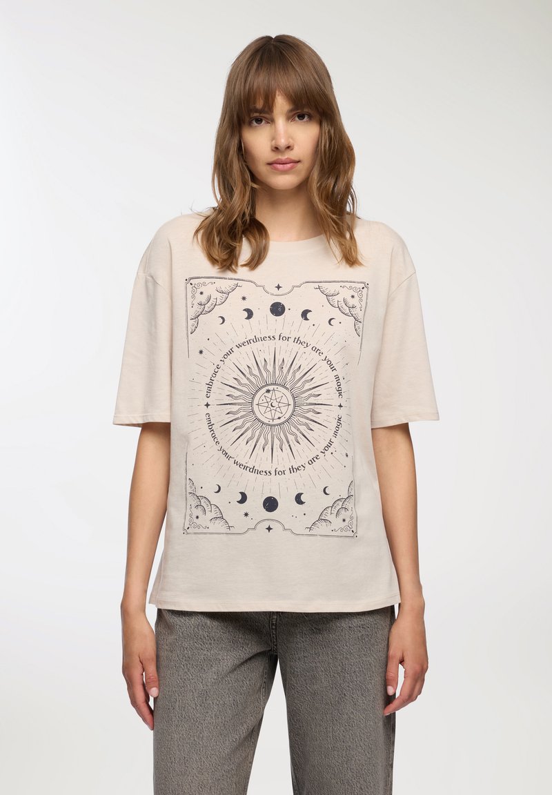 T-shirt in cotone beige con maniche corte, caratterizzato da un design grafico celestiale nero e testo al centro. Vestibilità morbida e rilassata, texture morbida.