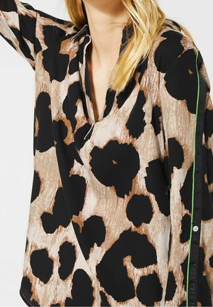 Femme portant une chemise ample marron et noire avec un grand imprimé floral abstrait et un détail de rayure verte sur la manche.