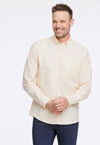 Ljusgul skjorta med knappar framtill, gjord av ett mjukt tyg, med en bröstficka och långärmad design. Kombinerad med mörkblå byxor.