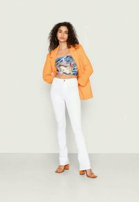 Blazer laranja, top cropped gráfico em azul e rosa, jeans brancos boca de sino e sandálias de salto laranja. Tecido suave e cores vibrantes.