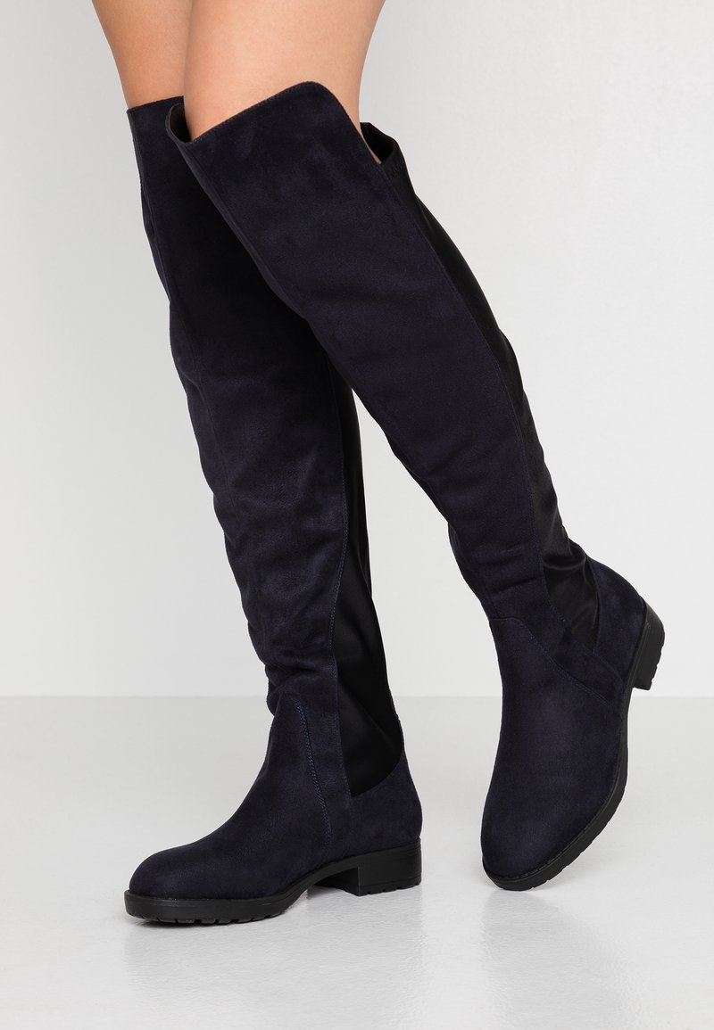 Jambes portant de hautes bottes noires en daim au-dessus du genou avec des talons bas sur un sol blanc et un fond clair.