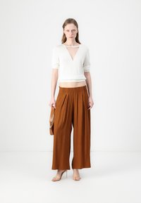 Witte korte mouwen top met een diepe V-hals, gecombineerd met brede bruine geplooide broeken. Model houdt een bruine tas vast en draagt transparante hakken.
