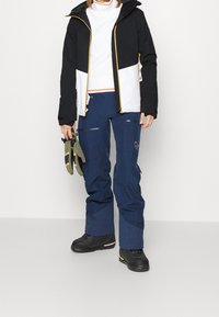 Veste de ski en noir et blanc avec des accents jaunes, associée à un pantalon bleu marine. Comprend des fermetures éclair, des poches et des bottes de ski noires. Gants tenus à la main.