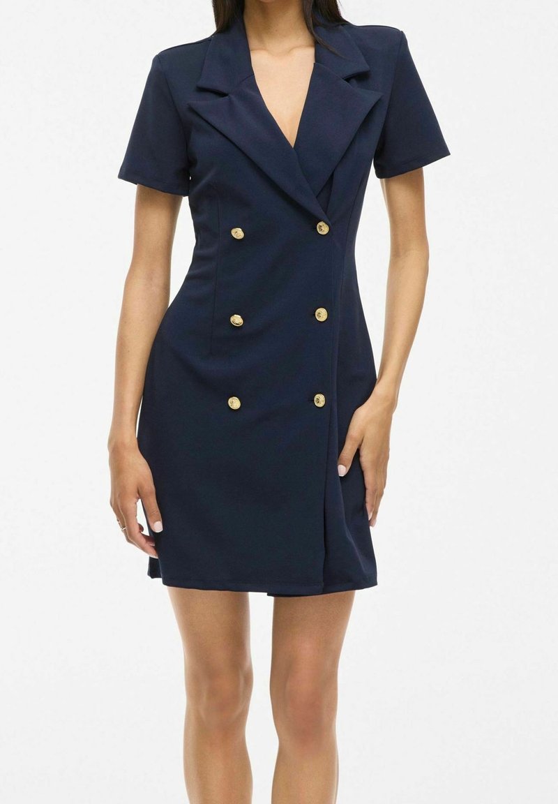Femme portant une robe-blazer bleu marine à double boutonnage, manches courtes et boutons dorés, debout avec une main touchant la robe.