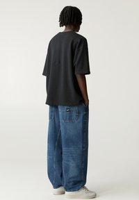 T-shirt en coton noir surdimensionné et jean en denim bleu ample. Le t-shirt présente un logo discret au dos ; le jean a de grandes poches arrière. Look décontracté.