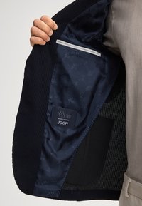 Blazer blu scuro con una texture a costine, dotato di una fodera interna in satin con un design a motivi e una tasca laterale per riporre oggetti.