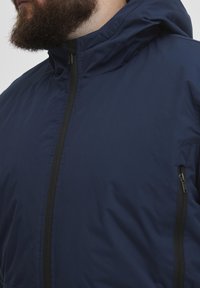 Jūros mėlynos spalvos lengvas švarkas su užtrauktuku, reguliuojamu gobtuvu ir dviem šoninėmis kišenėmis su juodais užtrauktukais. Lygus audinio paviršius.
