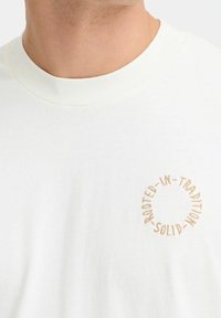 T-shirt en coton blanc avec un col rond. Présente une broderie circulaire beige avec les mots "EN-RACINÉ-DANS-LA-TRADITION SOLIDE" tout le long du périmètre.