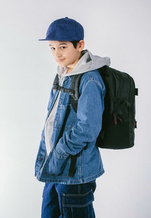 Schwarzer Rucksack mit verstellbaren Trägern an einer Person, die eine blaue Jeansjacke, einen grauen Kapuzenpullover und eine marineblaue Mütze trägt, kombiniert mit Cargo-Hosen.