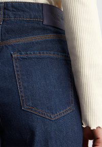 Tamno plave traperice s narančastim šavovima, s stražnjim džepom i kožnom oznakom s utisnutim "Marco O'Polo DENIM."