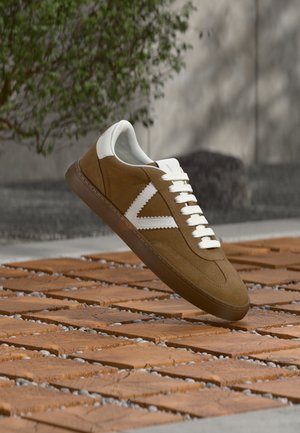 Brązowy zamszowy sneaker z białymi akcentami, sznurowany, z gumową podeszwą i teksturowanymi detalami. Na tle drewnianej powierzchni.