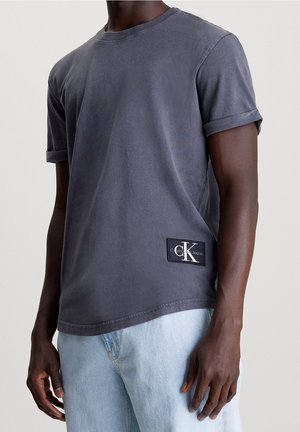 Mand iført mørkegrå T-shirt med korte ærmer og optrukne manchetter samt Calvin Klein-logo patch, kombineret med lyseblå jeans.