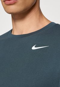 Tumman teal-pohjainen puuvillapaita, jossa on pyöreä kaula-aukko ja lyhyet hihat. Rintamuksessa on valkoinen Nike-logo. Sujuva rakenne.