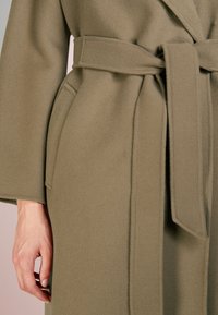 Manteau vert olive en tissu doux et texturé. Présente un design enroulé, une ceinture à la taille et des poches latérales invisibles.