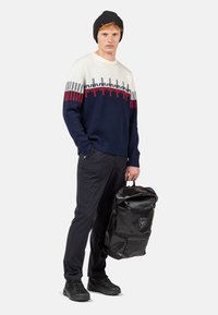 Marineblauer und cremefarbener Strickpullover mit rot-blauem Design, kombiniert mit einem schwarzen wasserdichten Rucksack, schwarzen Hosen und robusten schwarzen Schuhen.