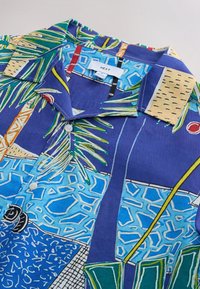 Chemise à manches courtes en coton bleu avec un motif tropical comprenant des palmiers, des formes géométriques et des accents contrastants beiges.