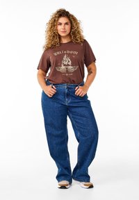 Smeđa grafička majica s dizajnom plamena i krila uparena s širokim plavim trapericama. Outfit je upotpunjen bijelim tenisicama.