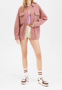 Przesadnie duża różowa dżinsowa kurtka z przednimi kieszeniami, z zapięciem na guziki, noszona na kwiecistym topie i postrzępionych białych szortach. Brązowe i białe sneakersy.