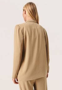 Beige bunda s dlouhými rukávy a měkkou texturou. Má klasický límec a jemné plisované detaily na ramenou. Hladký povrch, nezdobená zadní strana.
