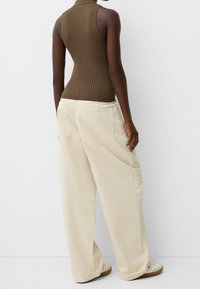 Personne portant un haut sans manches en maille marron, un pantalon ample de couleur crème avec des poches à l'arrière, et des baskets blanches, se tenant contre un mur uni.