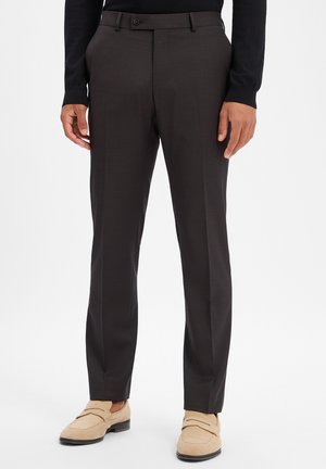 Mann trägt dunkelbraune, maßgeschneiderte Hose und beige Wildleder-Loafer, steht vor einem schlichten weißen Hintergrund.