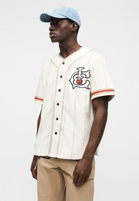 Κοντομάνικο μπέιζμπολ πουκάμισο σε κρεμ χρώμα με μαύρα κουμπιά, κεντημένα γραφικά και κόκκινες λεπτομέρειες. Συνδυάζεται με μπεζ παντελόνι cargo και τζιν καπέλο.