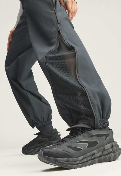 Person trägt schwarze Jogginghosen mit seitlichen Mesh-Einsätzen und schwarze klobige Sneakers mit grauen Akzenten, steht vor hellem Hintergrund.