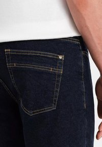 Jeans de mezclilla azul oscuro con costuras amarillas, diseño de bolsillo trasero con un remache metálico y tela texturizada. Detalle en primer plano del área de la cinturilla.