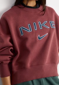 Osoba oblečená v vínovém Nike svetru s tmavě modrým logem a textem, spárovaná s tmavě zelenými kalhotami, zkrácenými u krku a pasu.