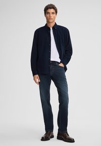 Camicia di velluto a coste blu navy sopra una maglietta bianca, abbinata a jeans di denim scuro e scarpe in pelle marrone. Modello in piedi contro uno sfondo neutro.