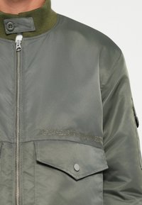 Giacca bomber verde con collo alto, chiusura a zip e tasche laterali. Il materiale ha una texture liscia. Presenta un logo stampato sul petto.