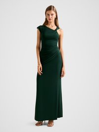 Long, dark green dress with a one-shoulder design, ruched texture, and subtle knot detail at the shoulder, paired with metallic heels.