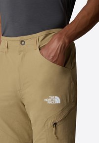 Pantalones de senderismo color caqui claro con un bolsillo lateral con cremallera, que cuentan con un logo blanco. La tela es suave y duradera, con un corte relajado.