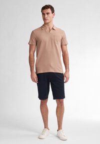 Polo tričko s krátkým rukávem, světle hnědé barvy, s náprsní kapsou, spárované s tmavě modrými šortkami. Model má na sobě bílé tenisky. Texturovaná látka.