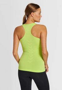 Woman with brown hair in a ponytail wearing a lime green racerback tank top and black leggings, facing sideways against a plain background.