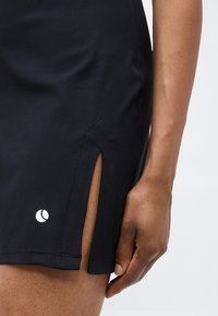 Primer plano de una falda negra con una abertura lateral, que muestra un logo parcial por encima del dobladillo y una mano apoyada junto a ella.