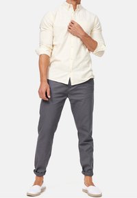 Hellbeige Langarmhemd mit Knopfverschluss und einer Brusttasche, kombiniert mit grauen hochgerollten Hosen und weißen Espadrilles.