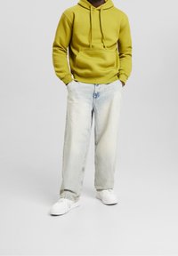 Sudadera amarilla con capucha y bolsillo frontal, combinada con unos vaqueros de pierna ancha de lavado claro y zapatillas blancas. Diseño sencillo y textura suave.