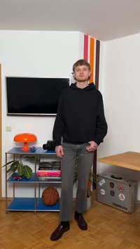 Tânăr cu tatuaje, purtând hanorac negru și blugi gri, stă într-o cameră cu un televizor, o lampă portocalie, un coș de baschet și o valiză argintie cu autocolante.