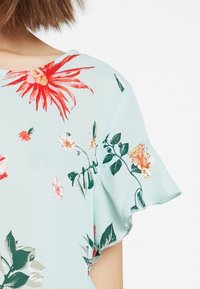 Světle modrá květinová blůza s červenými a růžovými květy, zelenými listy a volánovými krátkými rukávy. Hladká tkanina s jemnou texturou.