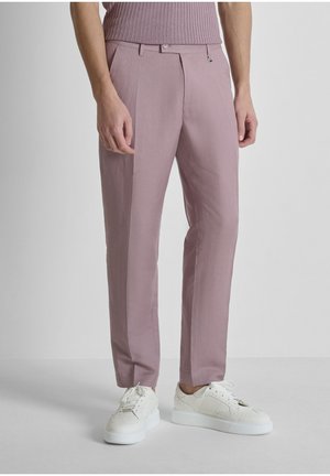 Hombre vistiendo pantalones a medida color malva, zapatillas blancas y una camisa de canalé malva metida por dentro, sobre un fondo gris liso.