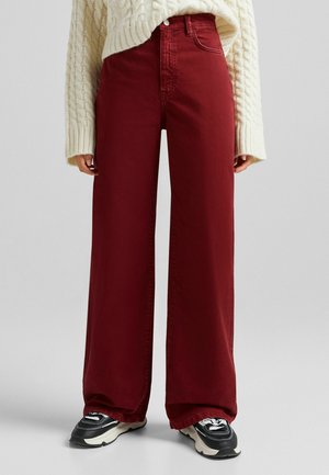 Personne portant un pantalon taille haute, jambes larges, de couleur rouge foncé, un pull crème en maille torsadée, et des baskets noires et blanches, debout sur un sol gris.