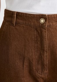 Des pantalons en coton marron présentent une coupe droite, des coutures visibles, un bouton doré à la taille et un tissu texturé, soulignant un design décontracté.