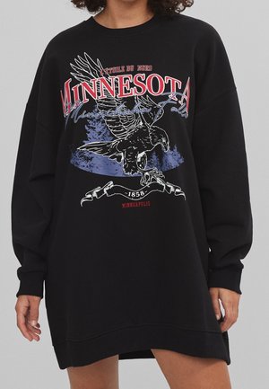 Personne portant un sweat-shirt noir oversize avec le texte "Minnesota", un graphisme d'aigle et des détails "Minneapolis 1858" à l'avant.